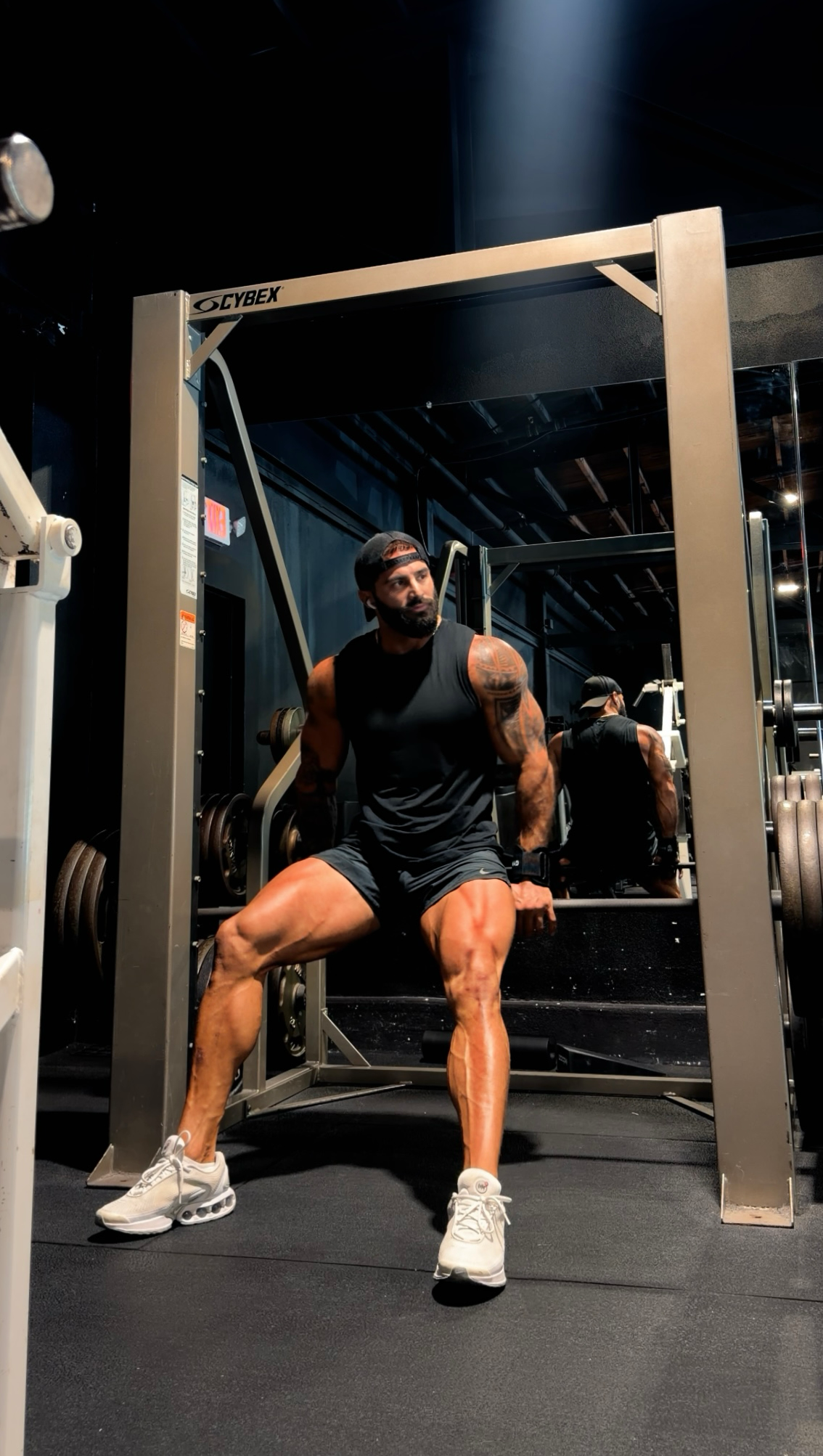 A muscular man wearing a black tank top, black shorts, and white sneakers sitting on a workout bench inside a gym, with a large Cybex workout machine behind him.