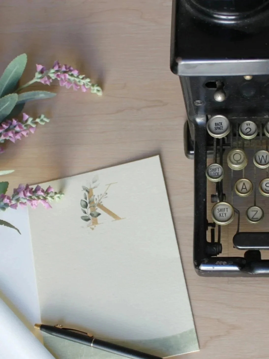 A vintage typewriter, a notebook with a letter 'K' decorated with green leaves and purple flowers, a black pen, and a sprig of purple flowers on a wooden surface.