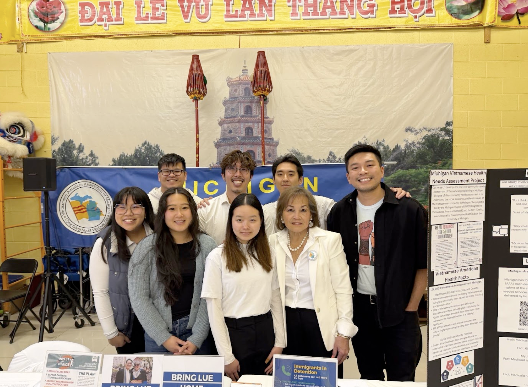 Group of people standing in front of a large banner with a picture of a pagoda and Vietnamese writing. The group includes young adults and an older woman, smiling at the camera, with informational posters on display nearby.
