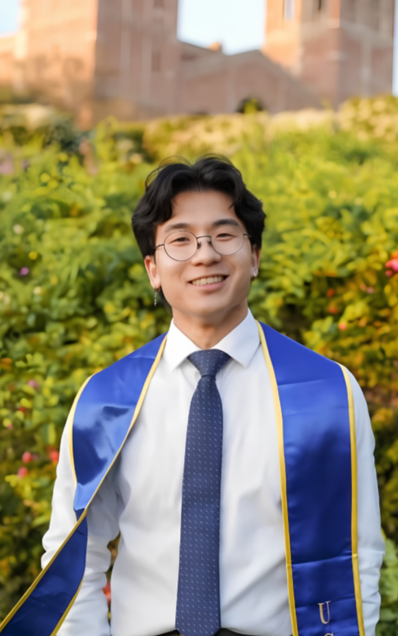 Young man in graduation attire with a blue sash, white shirt, and dark tie, smiling outdoors with greenery and a building in the background.