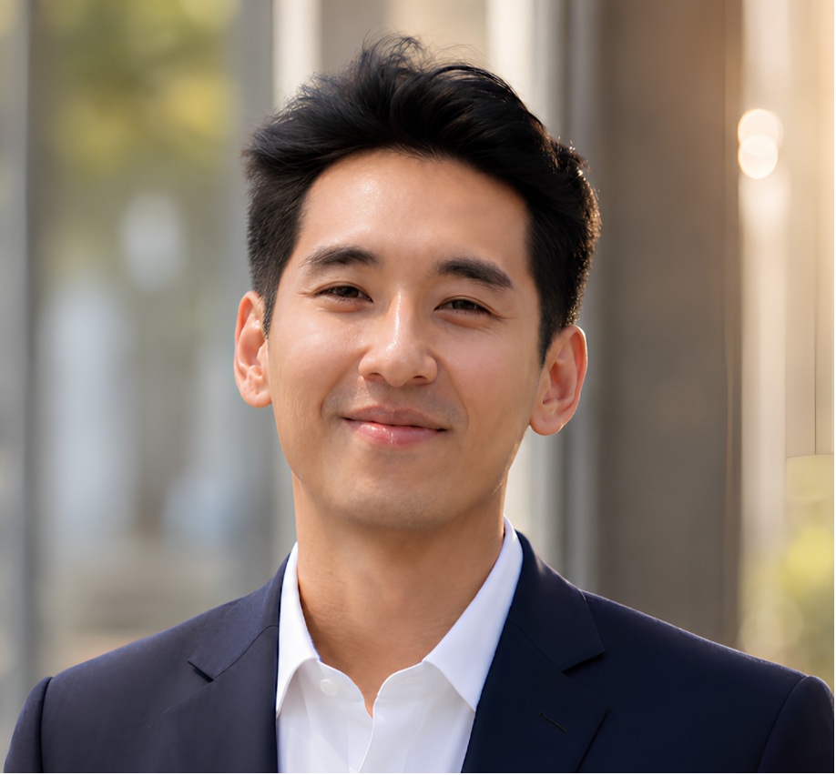 Young man in a navy suit and white shirt smiling, indoors with a blurred background of windows and sunlight.