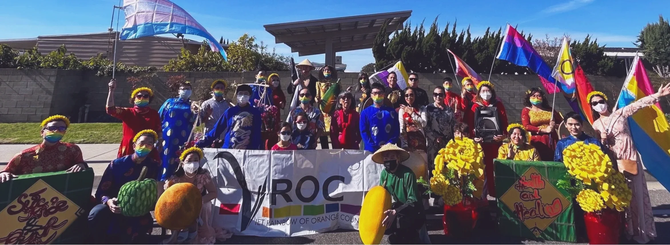 Group of people participating in a pride parade, holding rainbow flags, flowers, and signs, with some wearing rainbow-colored face masks and flower crowns, standing outdoors on a sunny day.