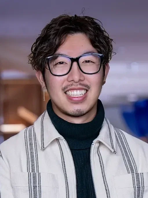 A young man with wavy dark hair, glasses, and facial hair, smiling and wearing a striped shirt over a black turtleneck, standing in an indoor setting.