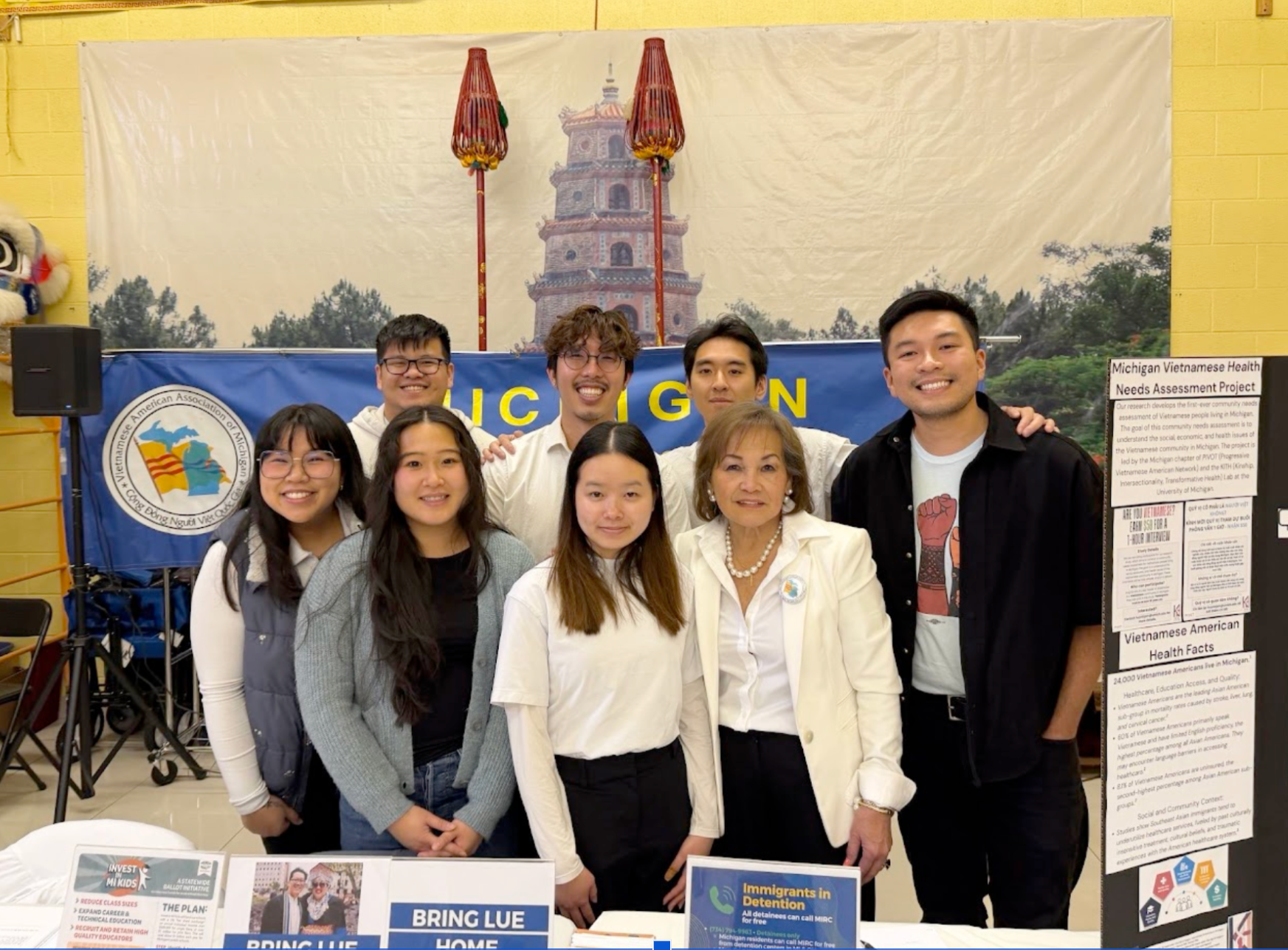 Group of eight people standing in front of a display booth for the Vietnamese American Health Needs Assessment Project, with a large backdrop of a pagoda and two red flags, and informational posters on the table and wall.