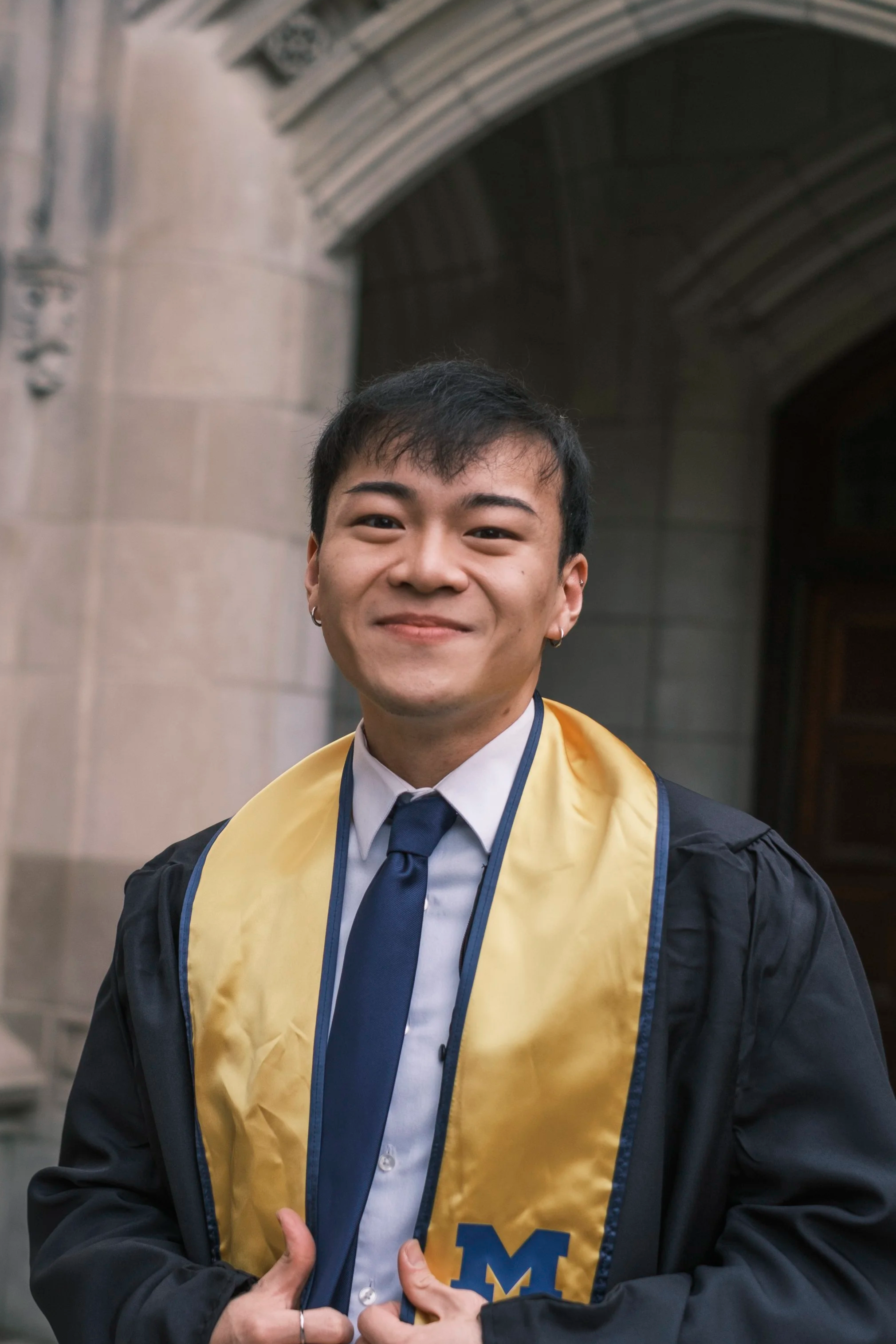Young man at graduation ceremony wearing a cap and gown, with a yellow stole and University of Michigan emblem, smiling.