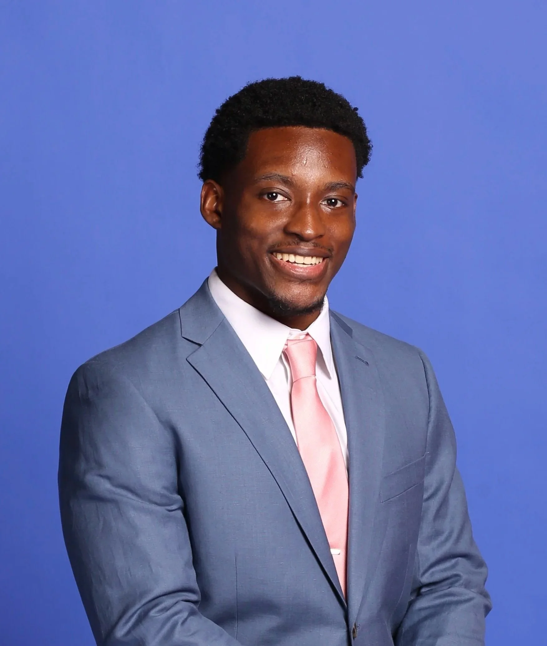 Young man with dark curly hair and a smile, wearing a light pink tie and a blue suit, standing against a solid blue background.