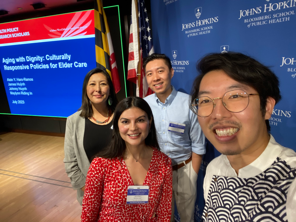 Group of four diverse people taking a selfie at a conference inside a room with a blue backdrop bearing the Johns Hopkins Bloomberg School of Public Health logo. A presentation slide titled 'Aging with Dignity: Culturally Responsive Policies for Elder Care' is visible on the left.