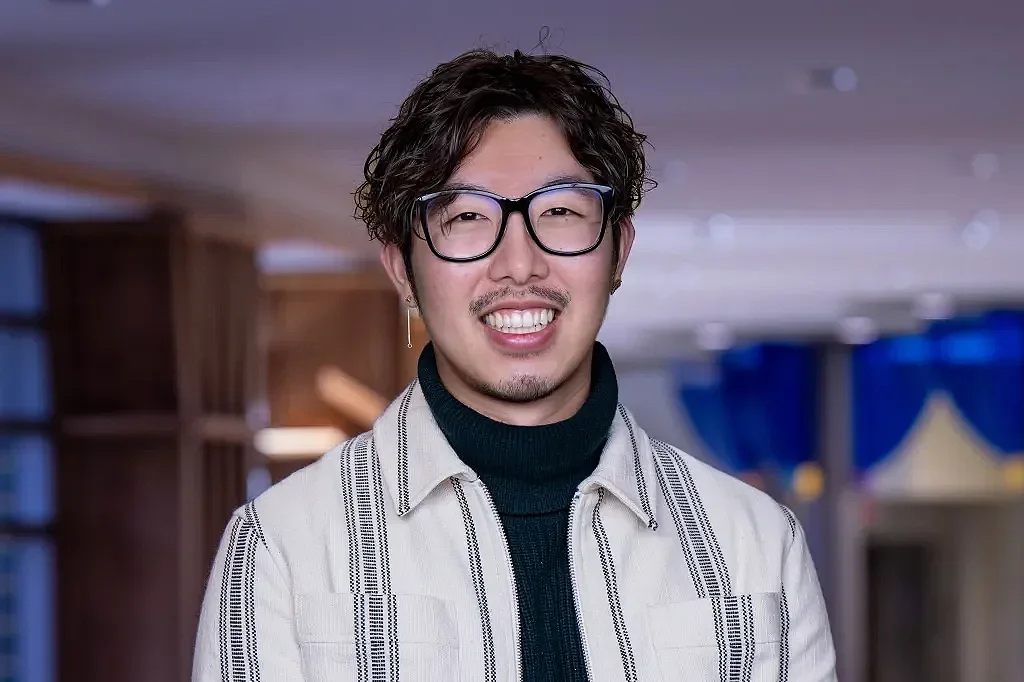A young man with curly hair, glasses, and an earring, smiling and winking in an indoor setting with blurred background.