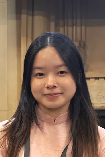 A woman with long black hair and light skin smiling slightly, wearing a pink traditional blouse, standing in front of a beige curtain and decorative wooden panel.