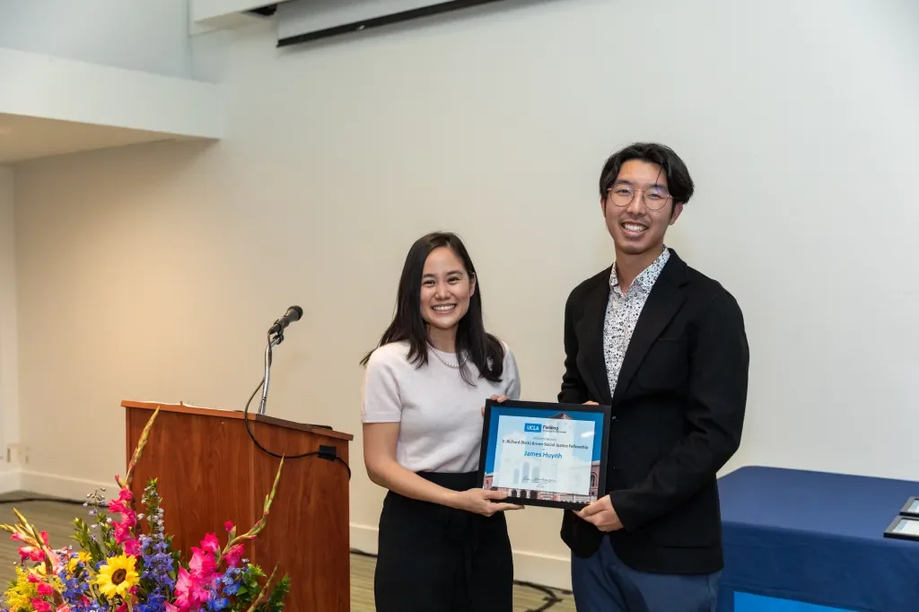 A man and woman standing together indoors, smiling at the camera. The woman is holding a framed certificate, and a table with colorful flowers and a microphone are visible nearby.
