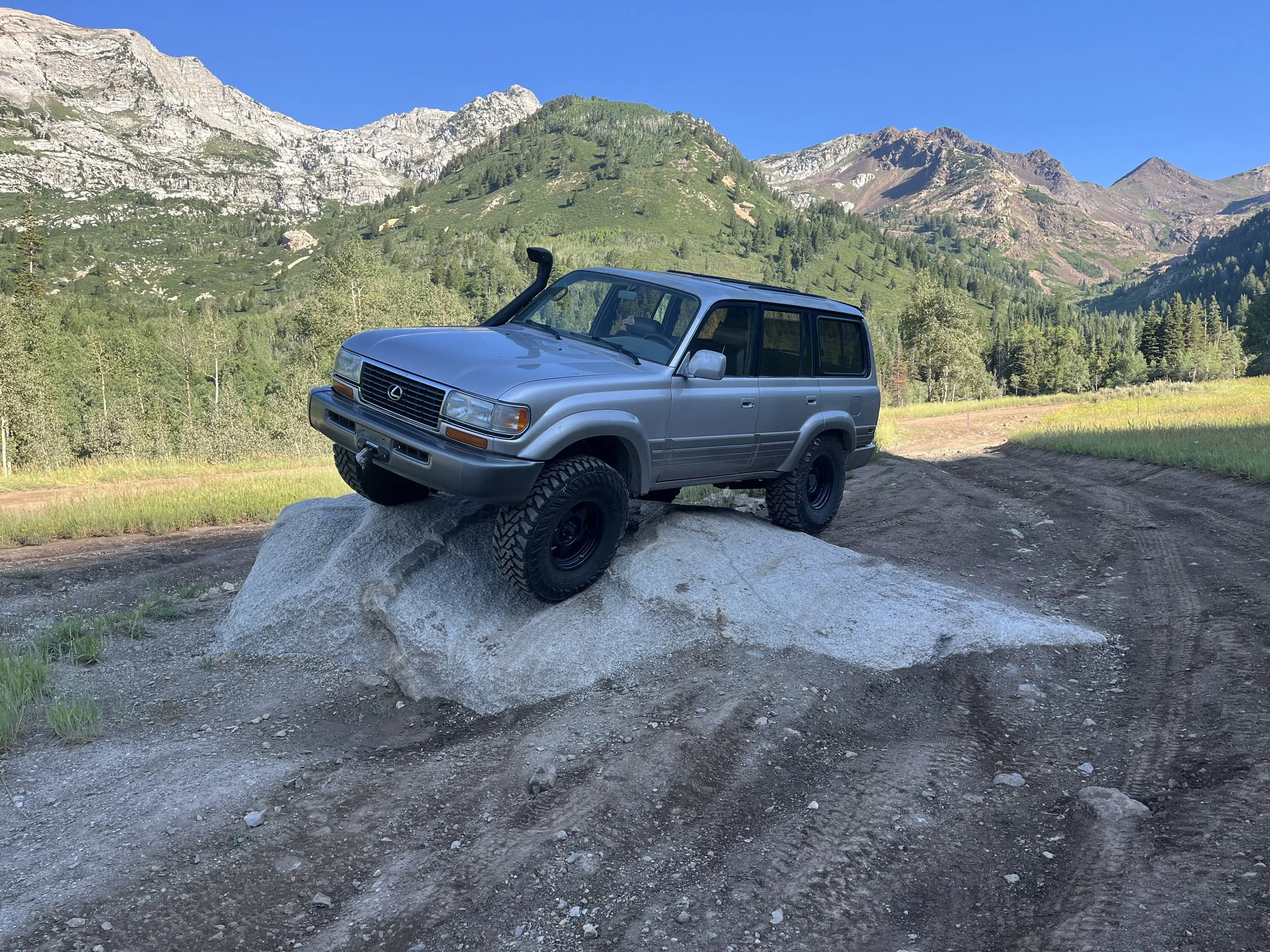 A silver SUV parked on a dirt trail in a mountainous area with green trees and rocky cliffs, under a clear blue sky.