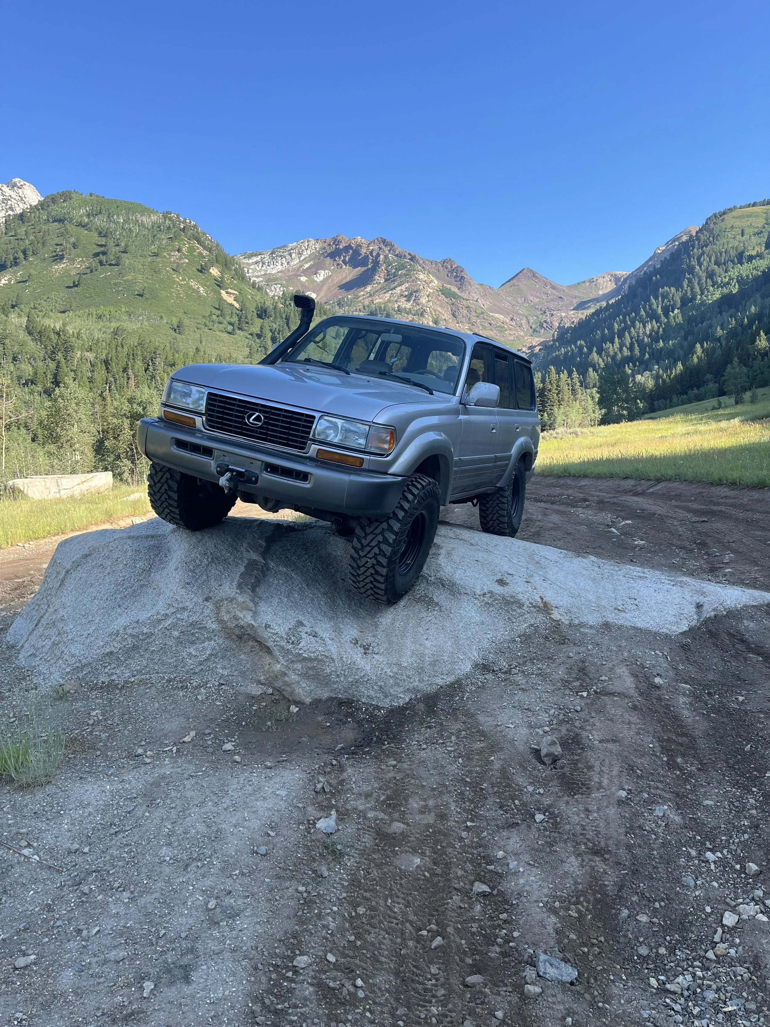 A silver Lexus SUV with large off-road tires positioned on a large rock in a mountainous area with green hills, rocky peaks, trees, and a clear blue sky in the background.