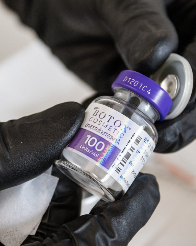 A close-up of gloved hands holding a vial of Botox Cosmetic, with purple cap and label indicating 100 units.