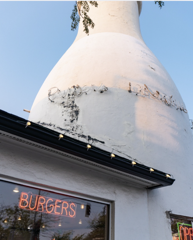Close-up of a white building with a chimney, a neon "BURGERS" sign in the window, and a small "OPEN" sign. The building has some paint wear and a wire sign attached to the chimney.