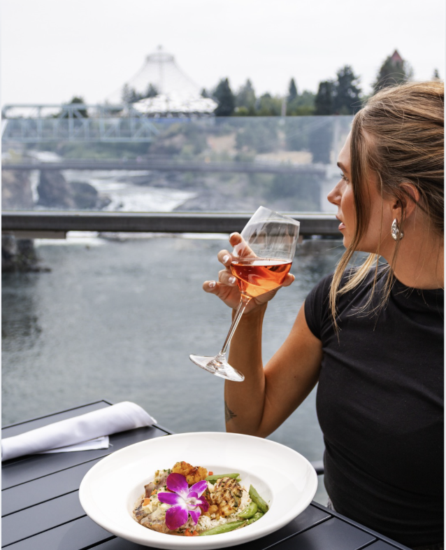 A woman with blonde hair, wearing a black top and earrings, is seated at an outdoor table, holding a glass of rosé wine near her face with a river and a bridge in the background. There is a plate of food with an orchid flower garnish on the table.