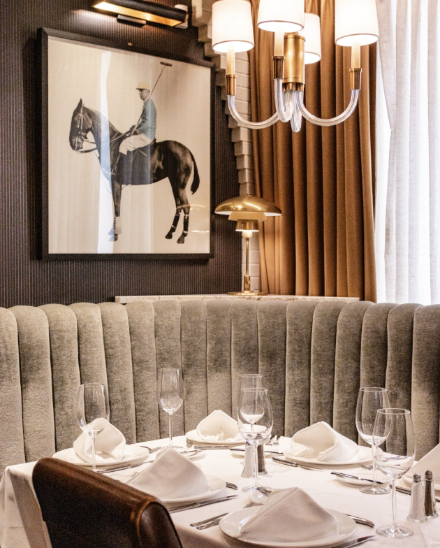 Elegant restaurant table set with white napkins, wine glasses, and silverware, surrounded by a curved gray upholstered banquette and brown leather chairs. A framed artwork of a person riding a horse hangs on the wall, with a modern chandelier and a gold desk lamp nearby, and beige curtains covering a window.