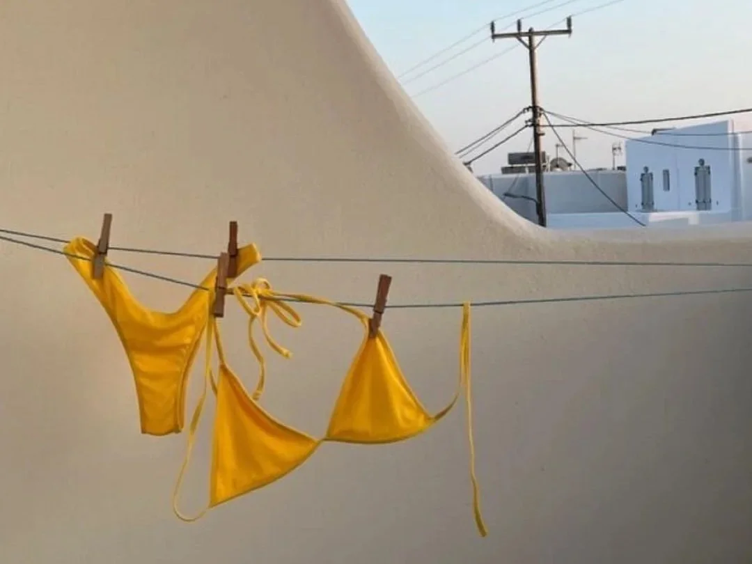 Yellow swimwear hanging on a clothesline with clothespins, against a white stucco wall and blue sky with electrical poles in the background.