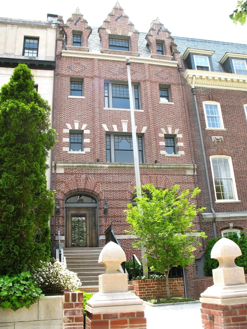 Front view of a historic brick residence with decorative gables, a staircase entrance, and surrounding greenery.