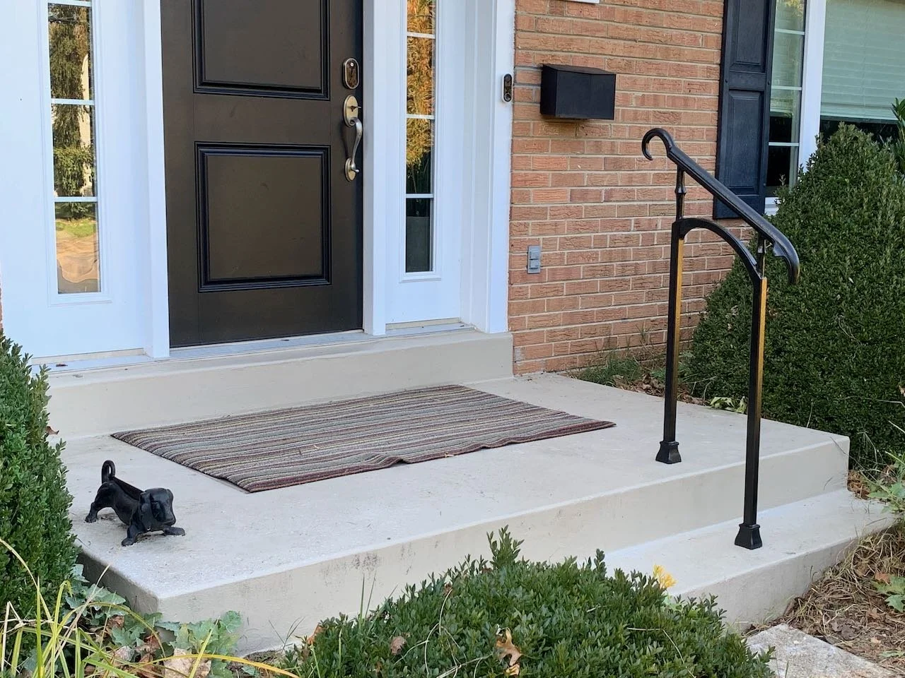 Front porch with black door, striped doormat, black handrail on steps, and bushes on sides.