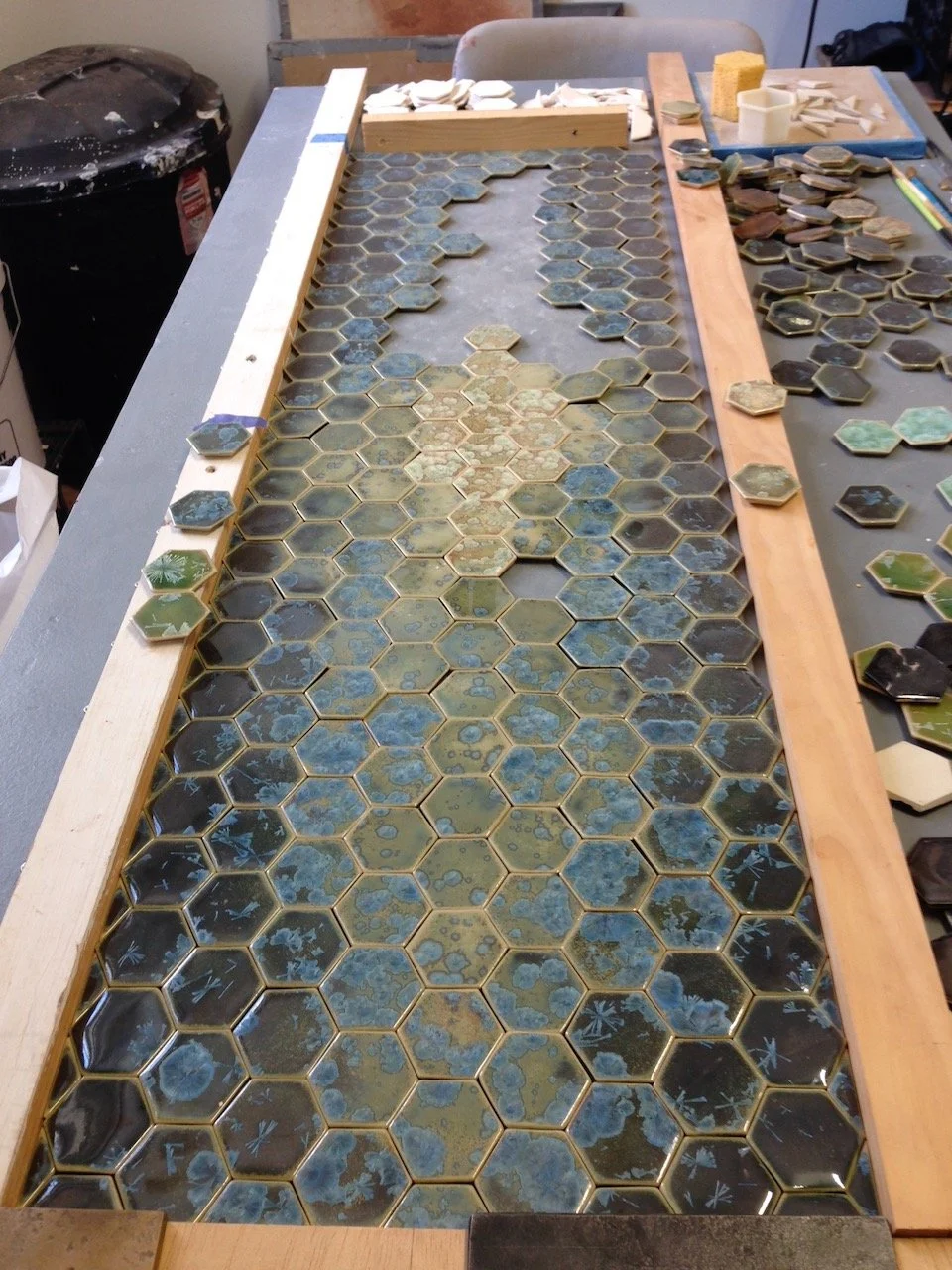 A work table with partially assembled hexagonal ceramic tiles in a mosaic project. The tiles are a mix of green, blue, and brown colors, with some still unglued and arranged in loose groups. There are wooden frames and spacers for alignment, and the workspace is cluttered with tools and supplies.