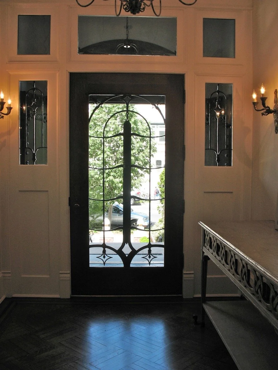 Interior view of a doorway with a decorative iron grille visible through the glass front door, flanked by wall-mounted black metal sconces with candle-style lights, and a glimpse of the street outside with parked cars and green trees.