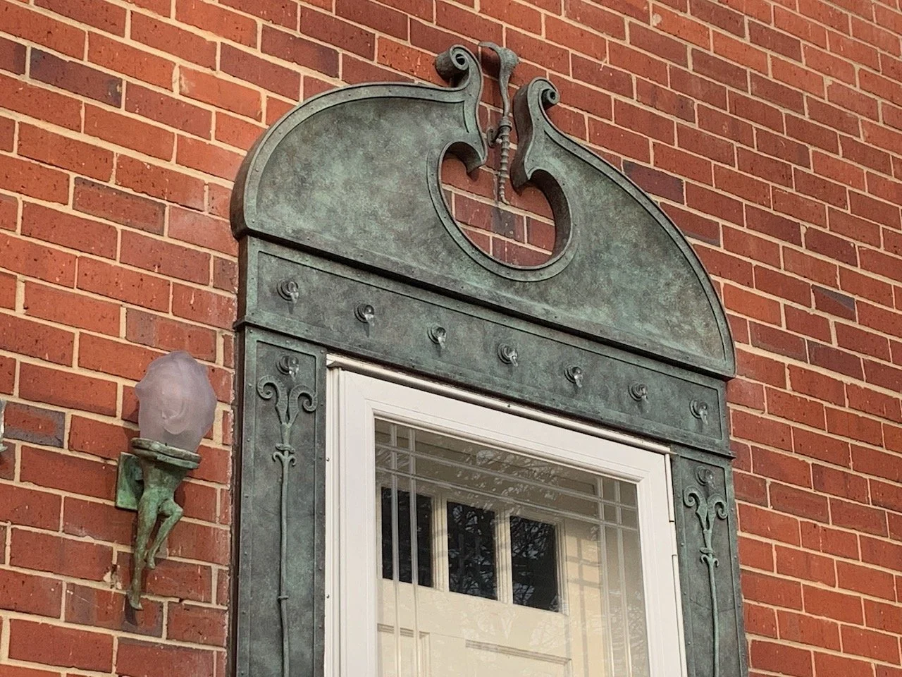 Close-up of a decorative bronze front door pediment  on a red brick wall, with an ornate light fixture on the left side.