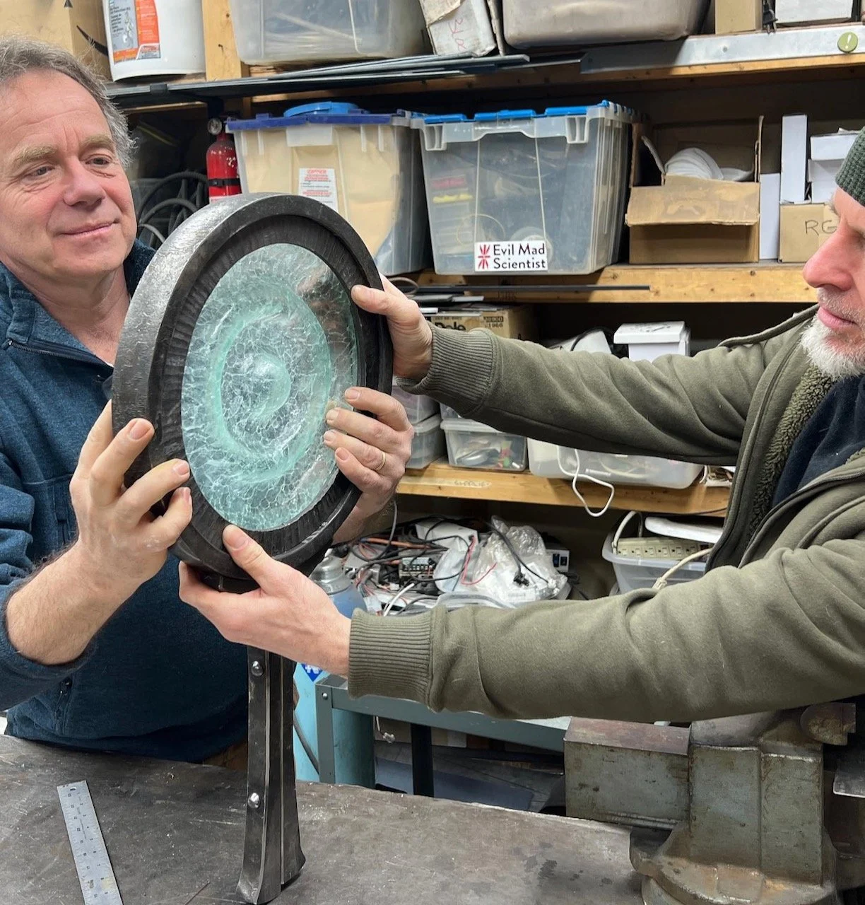 Two men hold a cast glass medallion in a forged iron frame, in a workshop filled with tools and storage bins.