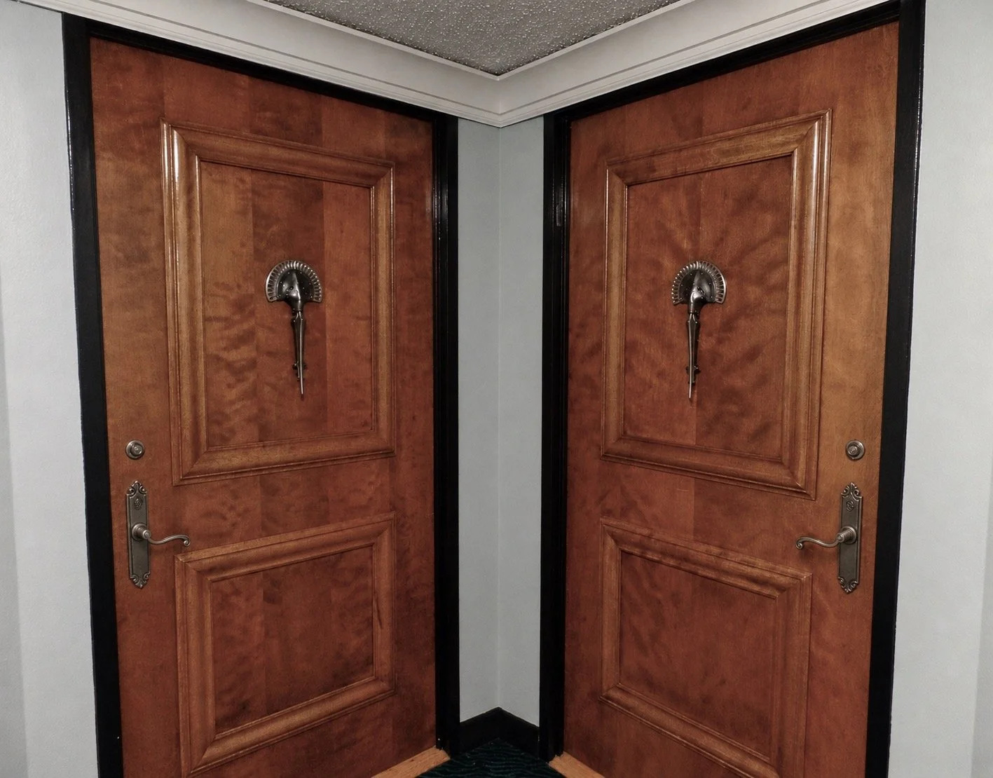 Pair of wooden doors with brass handles and custom designed door knockers in a corner, black trim, and white walls.