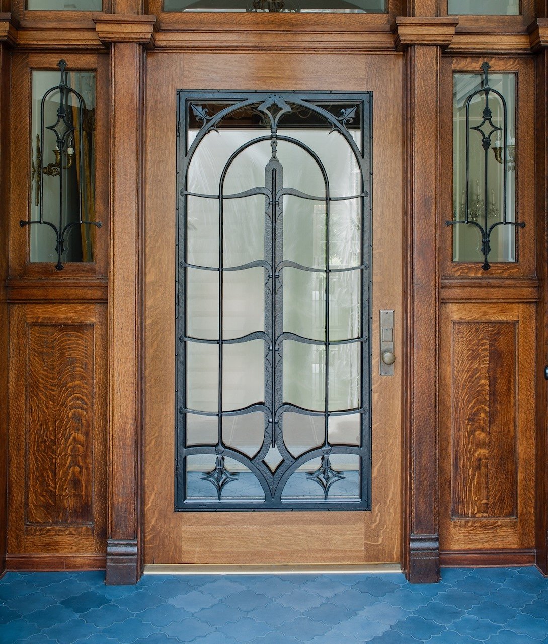 Wooden front door with decorative black wrought iron grille and frosted glass, flanked by wooden panels with small glass inserts and wrought iron accents.
