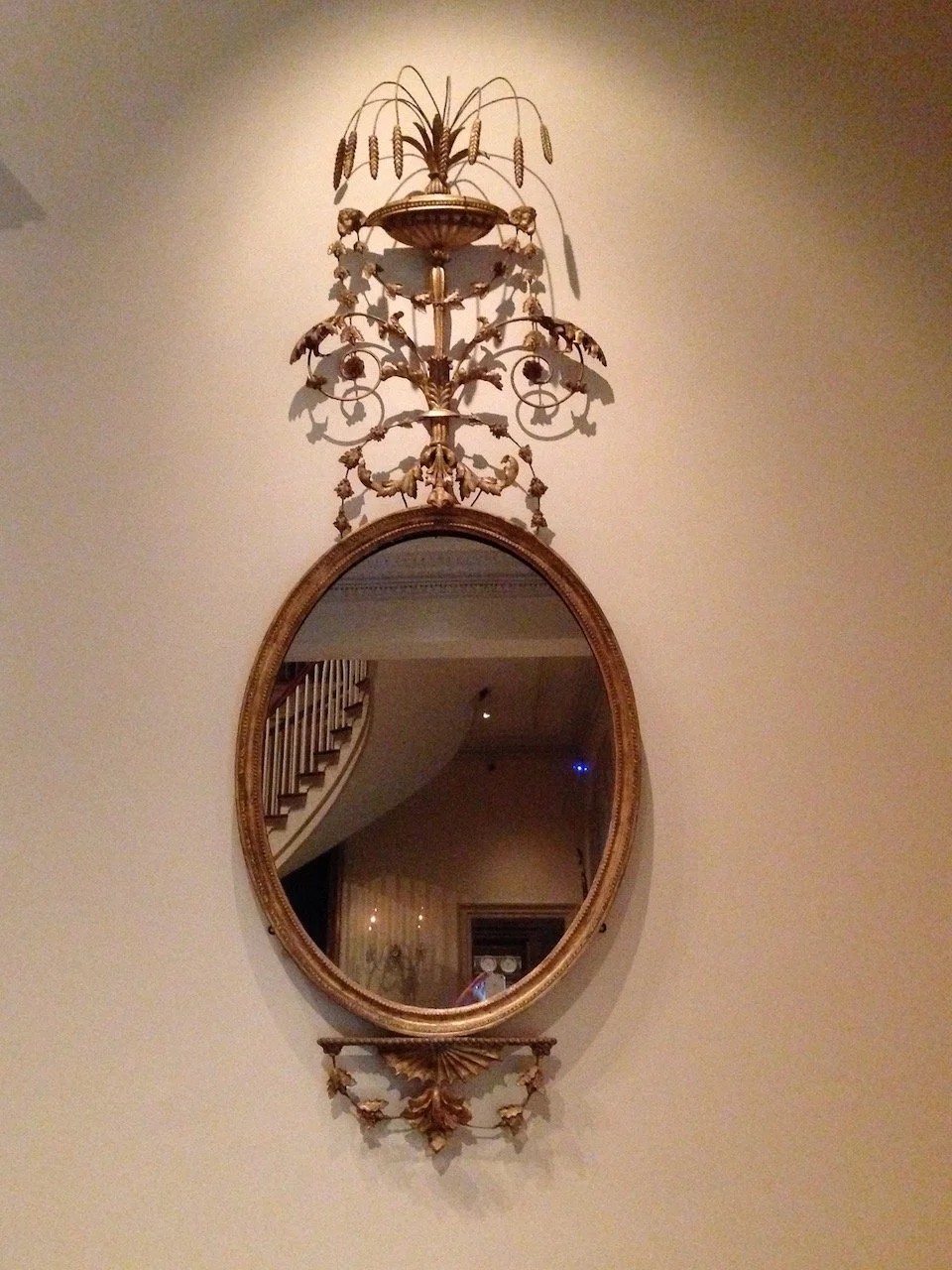 An antique oval mirror with a decorative gold frame hanging on a plain light-colored wall. Above the mirror is an ornate, vintage brass wall sconce with intricate floral and leaf designs, featuring a sculptural element at the top resembling a stylized plant or wheat motif.