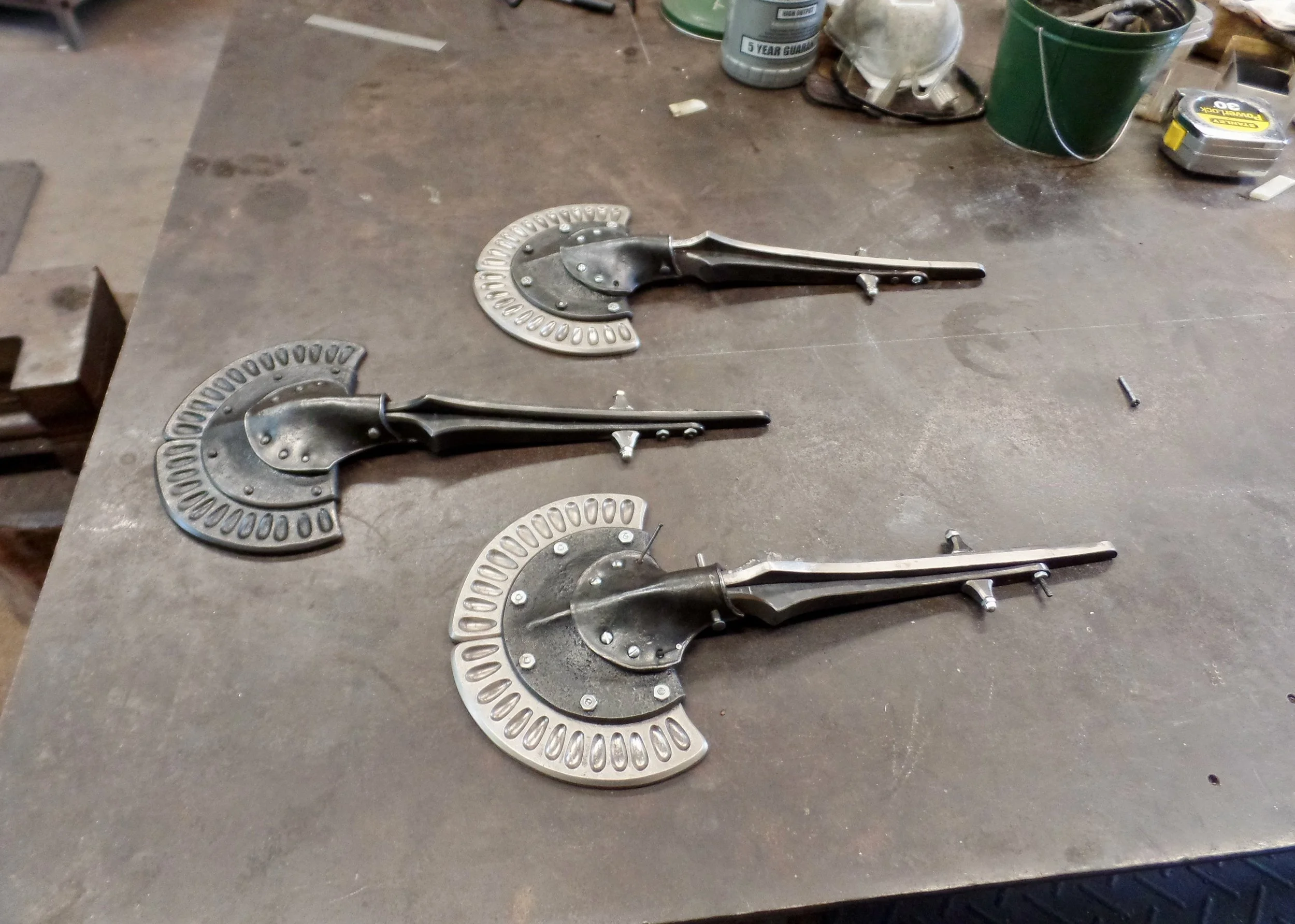 Three sculptural door knockers in forged metal placed on a workbench with tools and containers in the background.