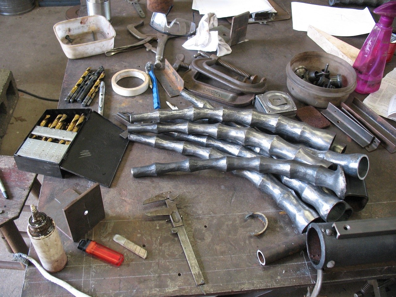 Worktable with metalworking tools, forged metal pipes, drill bits, clamps, and miscellaneous workshop items.