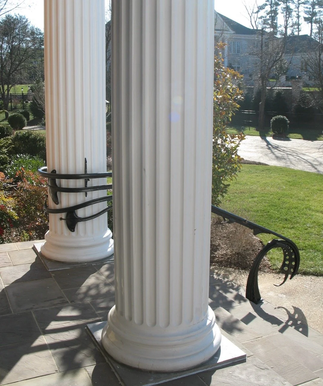 Close-up view of two large white classical columns with decorative bases, one of which has a black metal railing wrapped around it, situated outdoors on a patio. In the background, there is a grassy yard, bushes, trees, and a residential house.