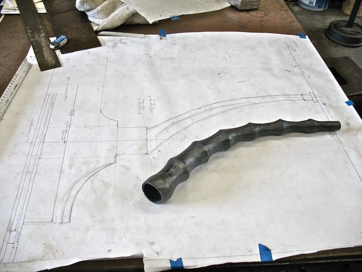 Metal hand-forged tool or pipe on a large technical drawing of a curved pipe or hose system.
