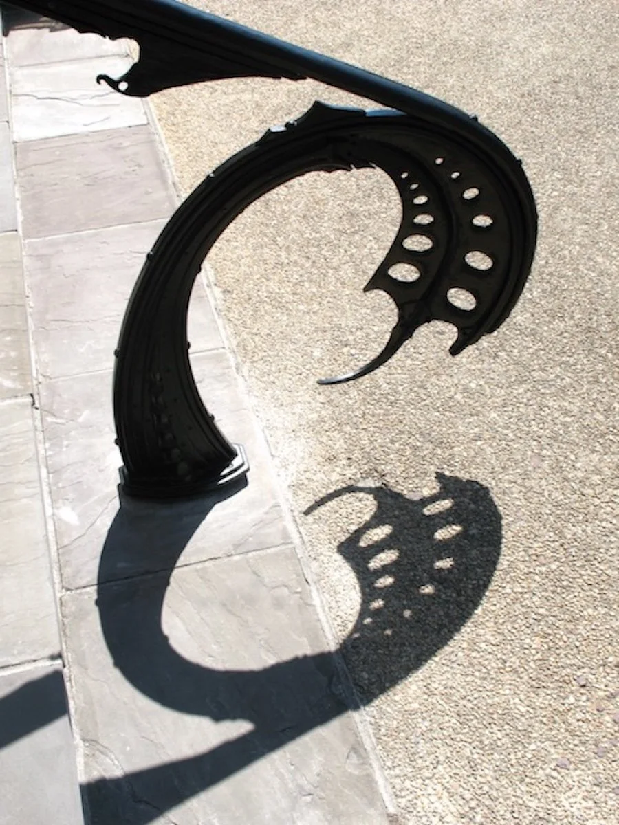 A decorative black metal handrail with a dragonfly design, casting a shadow on a tiled and gravel surface.