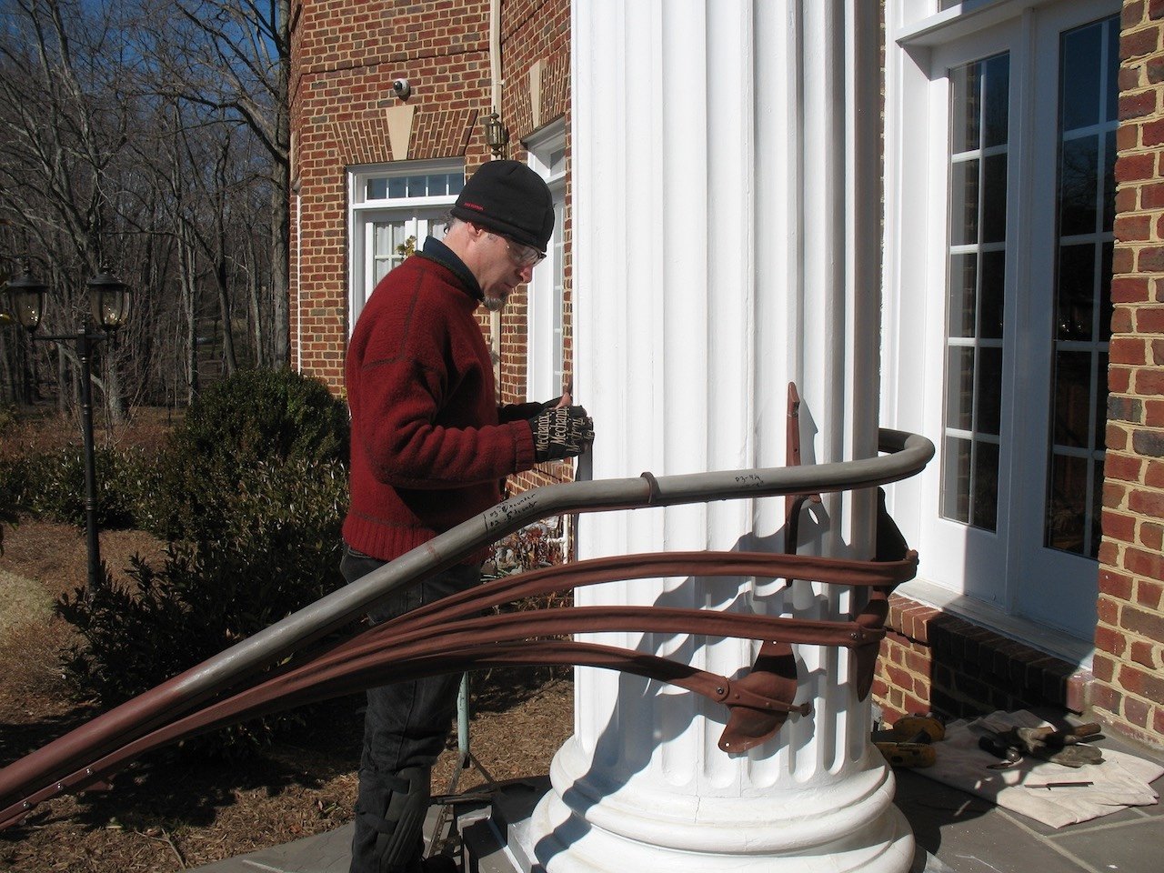 Man working on a porch railing outside a brick house with white pillars, wearing a black beanie, red sweater, and gloves.