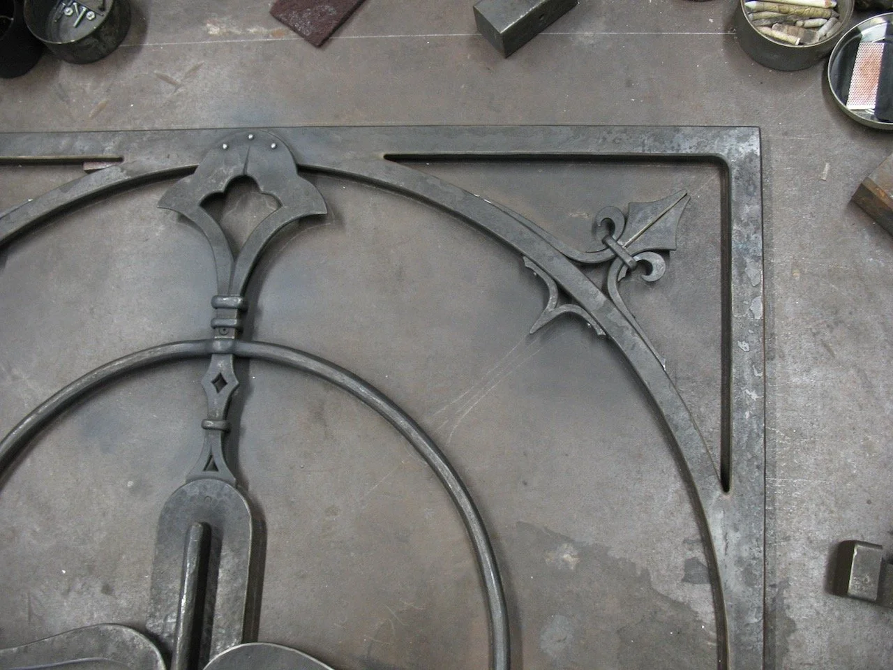 Close-up of a decorative metalwork piece, possibly part of a gate, featuring intricate design details and a curved shape on a workshop floor with tools around.