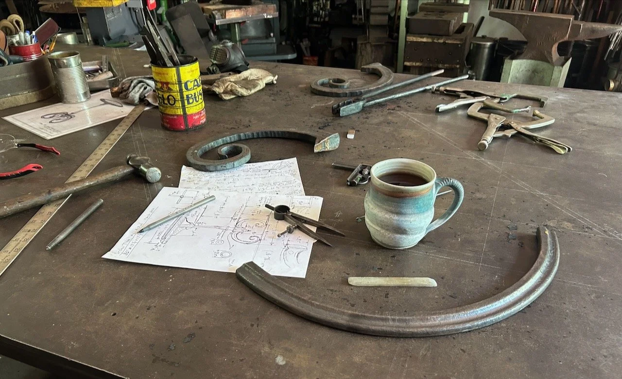 A cluttered workshop table with tools, a coffee mug filled with dark coffee, metal scrollwork, a ruler, pliers, sketches, and a can holding writing utensils.