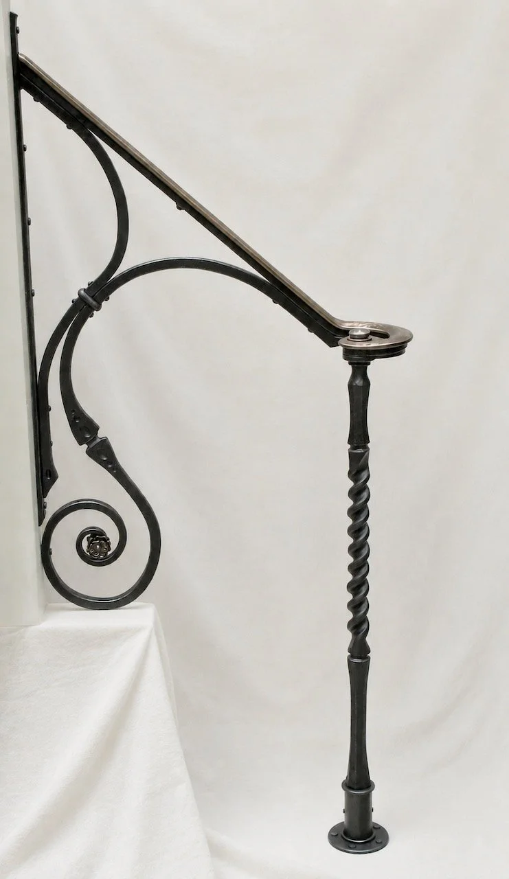 A forged iron handrail with a yellow bronze cap rail and a twisted iron newel post against a white backdrop