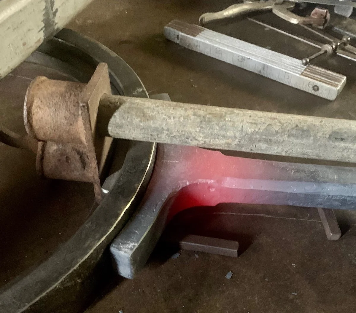 A rusty hydraulic press with a metal bar placed across its platform, surrounded by various metal tools, including a square ruler.