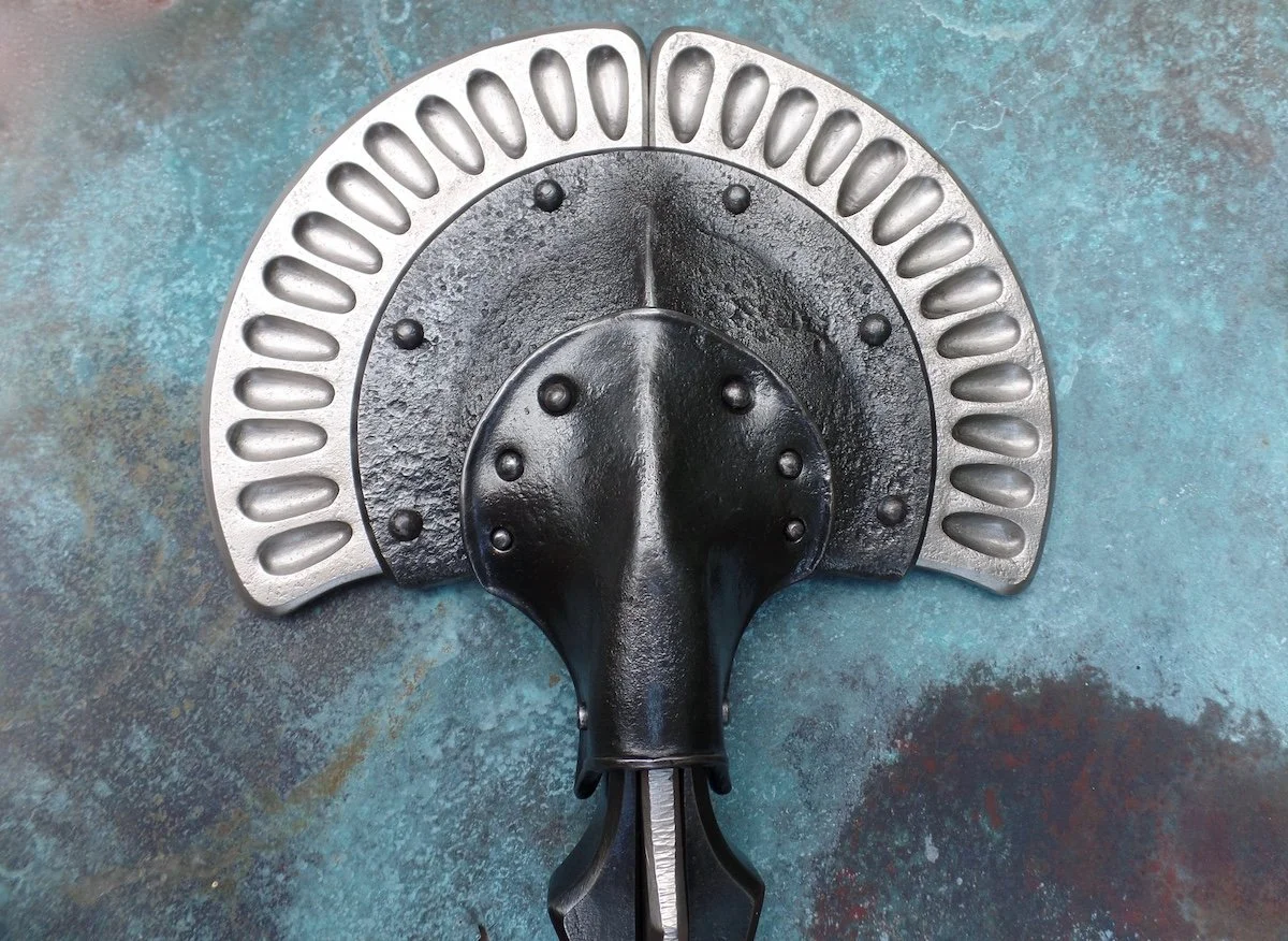 A black and silver sculptural forged metal door knocker placed on a blue-green patinated bronze surface.
