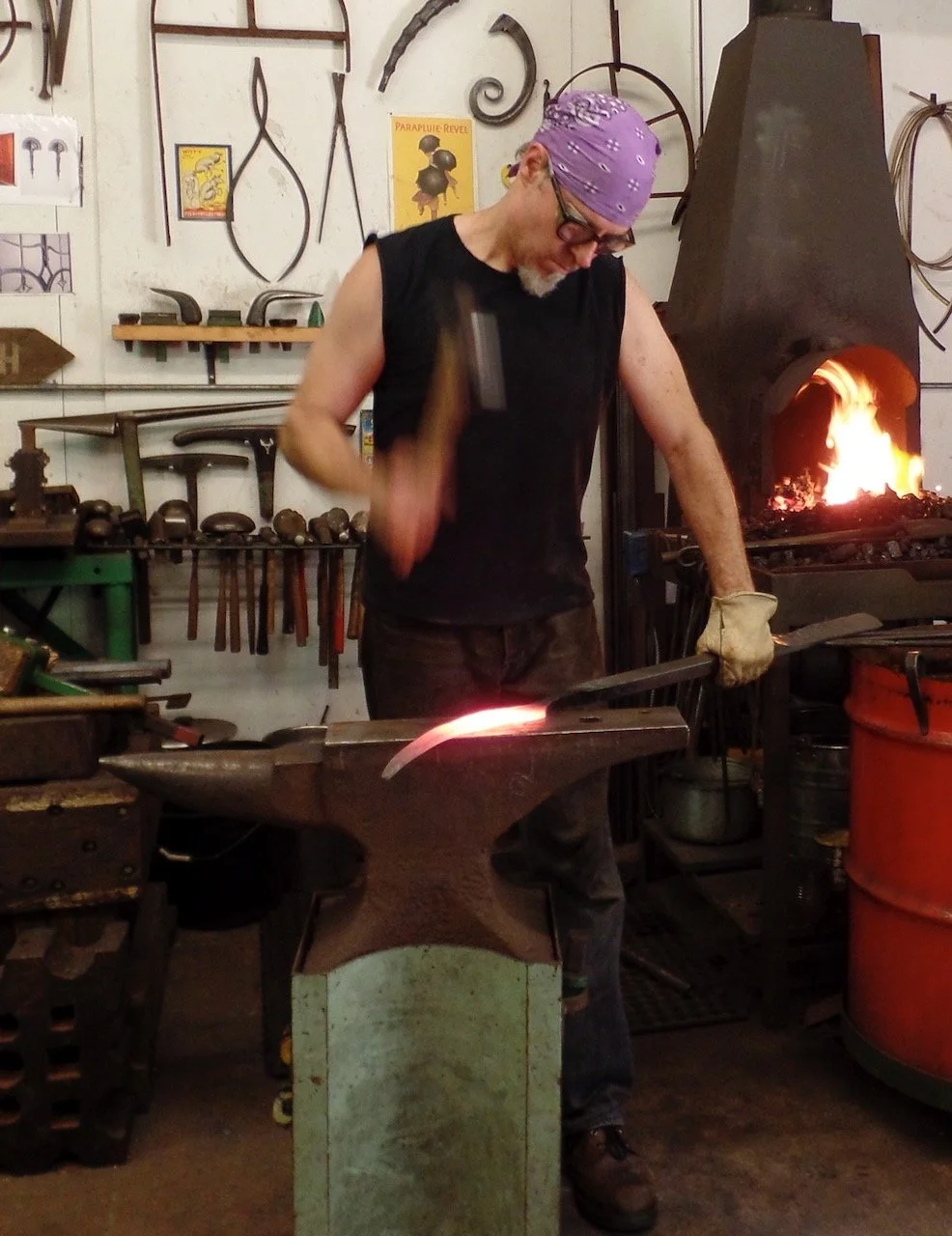 Blacksmith wearing glasses, purple bandana, and sleeveless shirt, hammering a glowing piece of metal on an anvil in a workshop with various tools and a forge with fire in the background.
