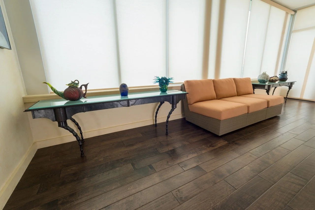 Living room with a peach-colored sofa, long glass-top console tables with decorative vases and plants, and large windows with blinds.