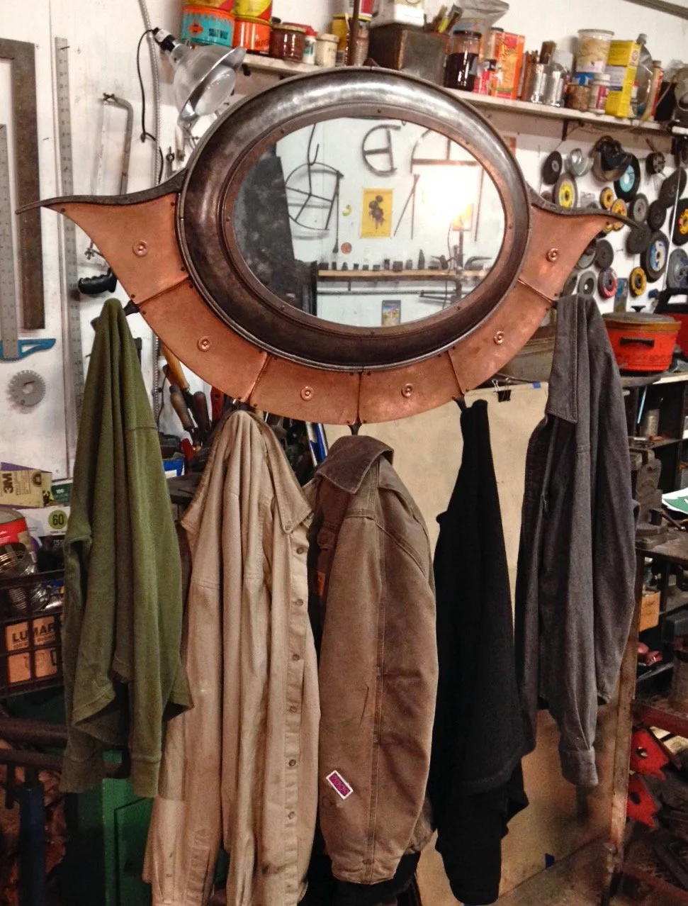 A garage wall with a large, vintage, bat-shaped mirror, with clothes hanging beneath it and shelves with tools and supplies in the background.