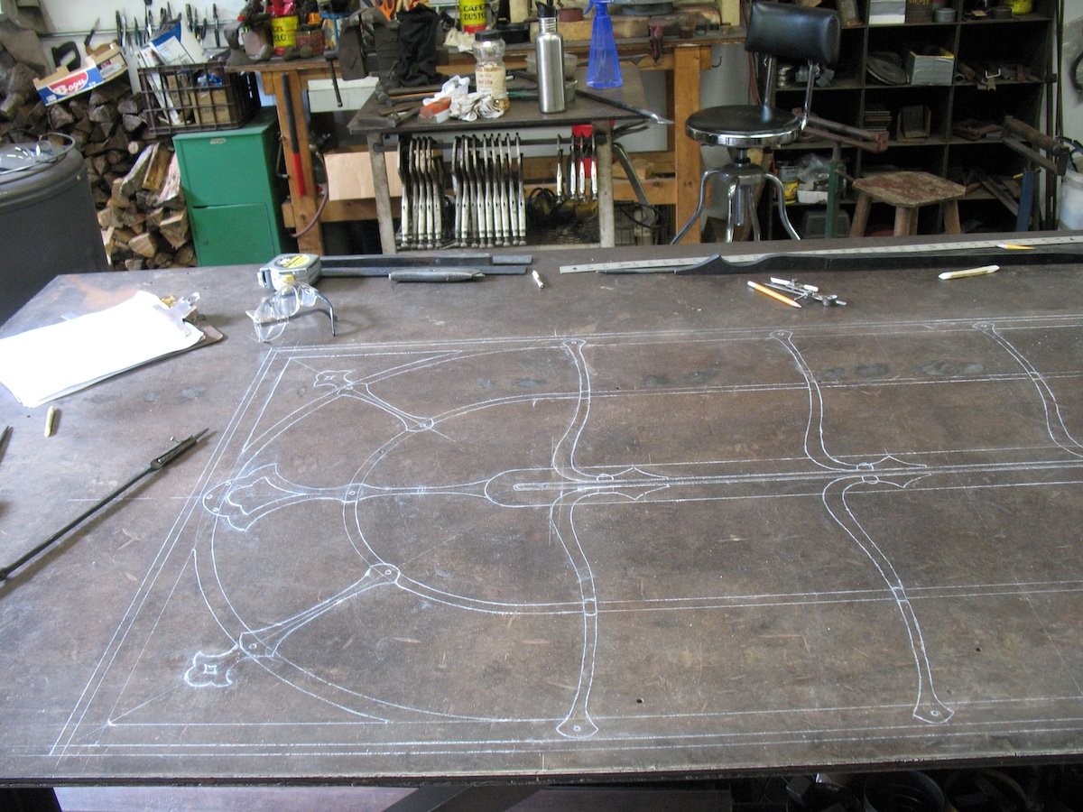 A blacksmith workshop with a large workbench that has a detailed design or blueprint drawn on its surface. The background contains various tools, equipment, and materials, including knives, a green storage cabinet, and stacks of firewood.