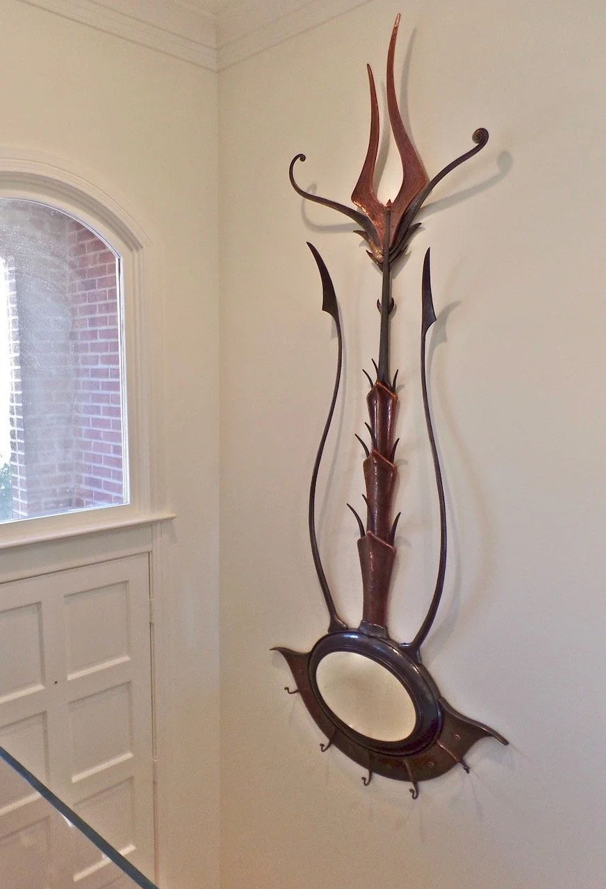 Decorative wall piece featuring curved iron and copper forgings, an oval mirror, and sculptural elements, mounted on a white wall near a window.