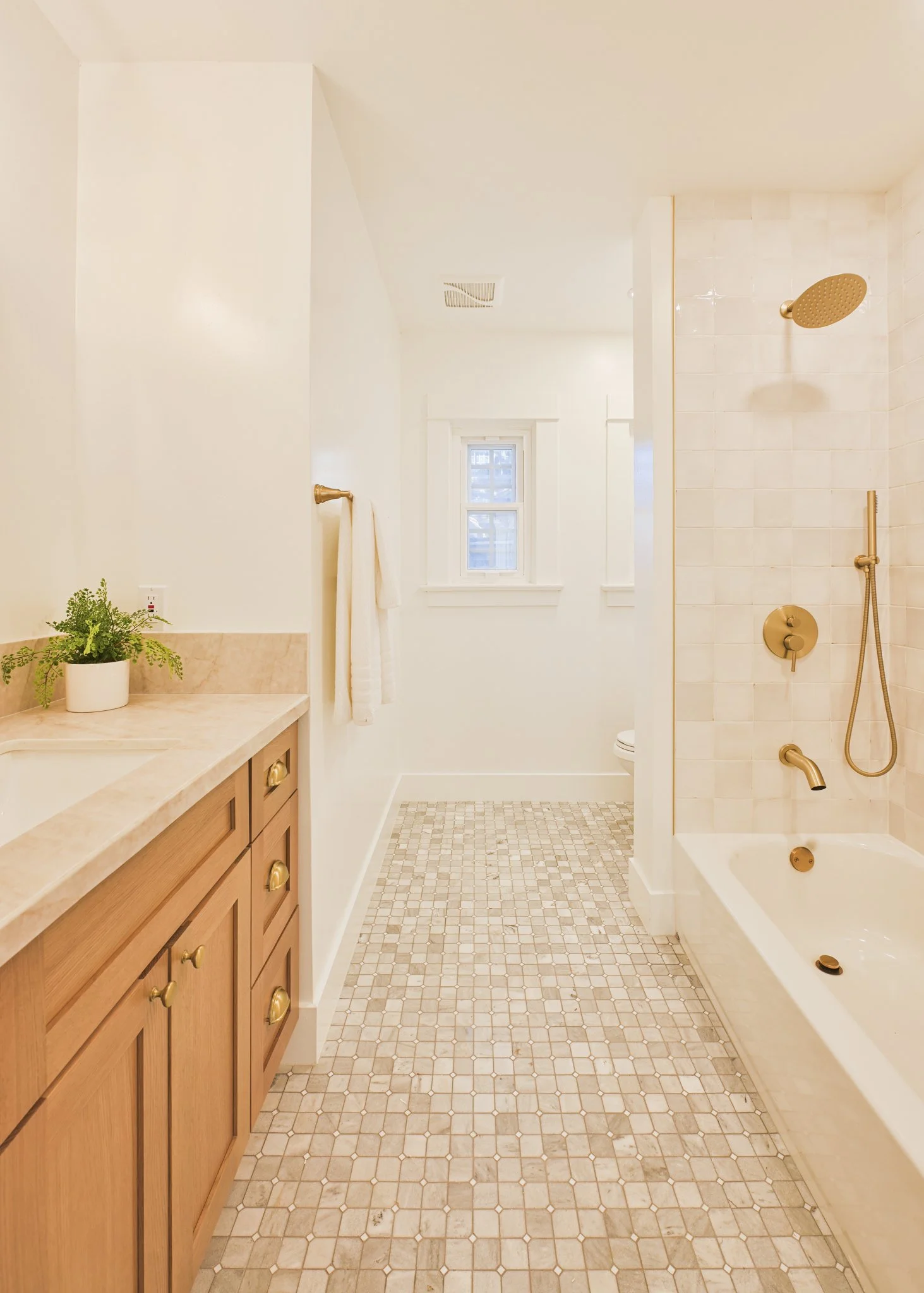 Bathroom with wooden vanity, beige countertop, potted plant, white walls, small window, and a separate shower area with gold fixtures and a bathtub.