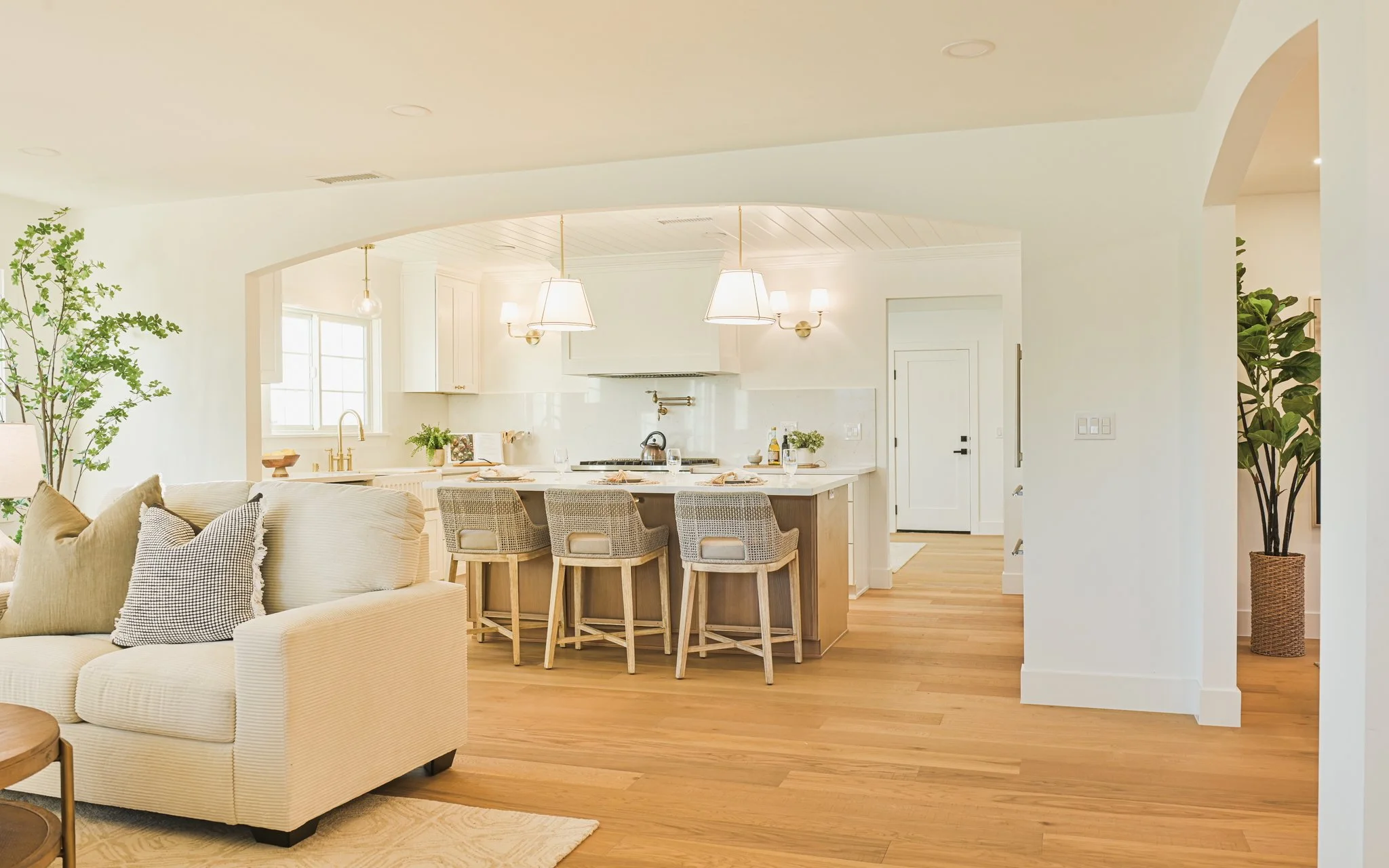 Open-concept living room and kitchen with neutral colors, wooden floors, and modern lighting fixtures, including a beige sofa with patterned pillows, a kitchen island with four stools, and tall green houseplants.