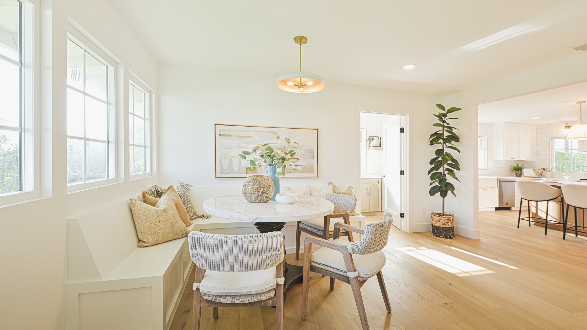 Bright living room with large windows, white walls, and wooden flooring. Features a marble-topped dining table with assorted chairs, a built-in bench with cushions, wall art, potted plants, and a view into a kitchen area.