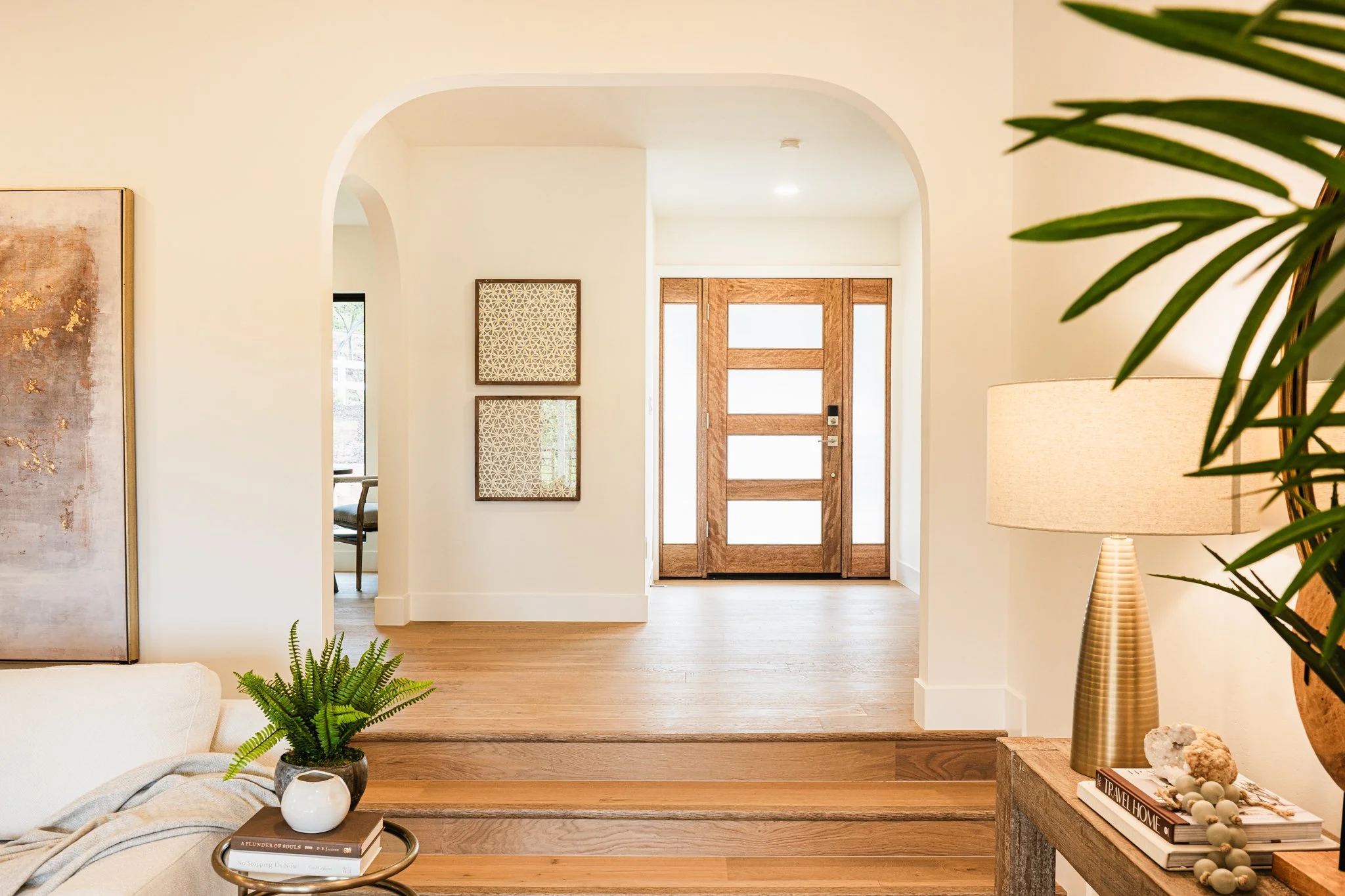 Corner of a living room with a table holding books, a white vase, a green potted plant, and a blanket. In the background, an entrance with a wooden door and wall art, and a segment of a small chair is visible through a doorway.